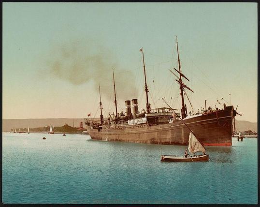 The image depicts a large steamship docked in the water, with smoke billowing from its stack indicating it's either arriving or departing. The ship has multiple masts and rigging that suggest an older style of seafaring vessel. In the foreground, there is a smaller sailboat carrying one person, which adds scale to the scene and provides context for human activity in relation to the large ship.

The water appears calm with several other small boats visible at various distances from the viewer's perspective. The background shows hazy skies over what seems like a coastal area or port city, where more ships can be seen on the horizon along with some structures that resemble lighthouses or signal towers.

This photograph is likely of historical significance as it captures an early 20th-century scene involving maritime activity in Egypt's Suez Canal. The image appears to have been colorized or altered for artistic effect and bears a vintage tone, indicating its age.