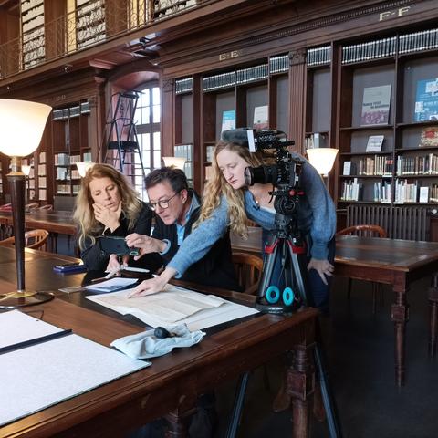 Dans une bibliothèque anciennes 3 personnes sont penchées sur un document ancien. Il y a une caméra à côté d'eux.
