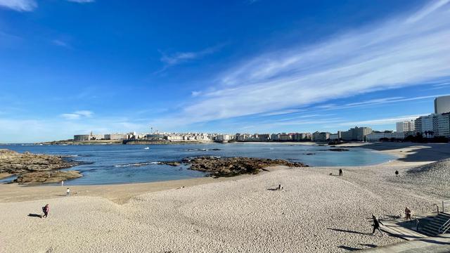A scenic coastal view featuring a sandy beach with sparsely populated areas, rocky formations, and calm waters. In the background, there are modern buildings and a blue sky with wispy clouds.