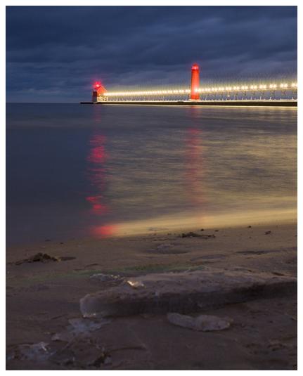 A wide view of a long concrete pier jutting out into Lake Michigan with a catwalk with lights on it along the pier. At the end are a red lighthouse and secondary building with red lights on top. A moody, cloudy sky looms overhead.