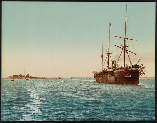 The image shows a large ship with multiple masts, sailing across the water. The vessel appears to be an older style of maritime transport, possibly from the late 19th or early 20th century, given its design and the presence of flags on its rigging which may indicate nationality or affiliation at that time period. It is cruising along what seems like a coastal area with land visible in the background across calm waters, likely partaking in trade or travel routes during this era.

The sky has a muted mix of light blue and soft yellow hues, suggesting either early morning or late afternoon lighting conditions. The water reflects these colors as it moves gently through the scene. There are several small boats scattered around near the shore, indicating activity within this coastal region. In the distance, there is another larger ship on the horizon line.

The text "Canal de Suez" and a date of approximately 1890-1910 can be seen at the bottom right corner, which implies that the photo might have been part of a series documenting various aspects or locations related to Egypt's Suez Canal during this period. The photograph carries an antique quality with its warm sepia tones typical for early colorized prints from around 1875-1904 using the photogravure process.

This image is indicative of historical maritime exploration and trade, showcasing a moment in time where such vessels played crucial roles in connecting continents vi [...]