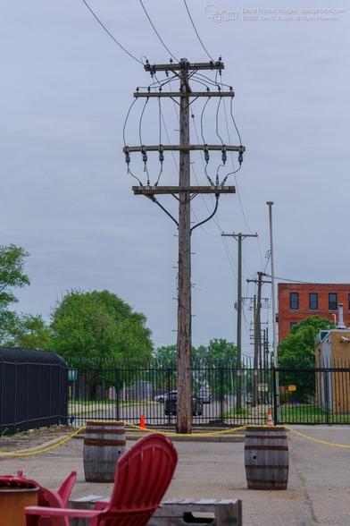 A photograph of a weathered wooden utility pole, supporting a network of electrical wires, in a paved area.  A black wrought-iron fence surrounds a parking lot or similar area, with a few parked cars visible beyond.  Red Adirondack chairs and wooden barrels are situated in the foreground, indicating an outdoor seating or gathering area.  The image shows an urban landscape with utility infrastructure and a hint of industrial activity in the background.
