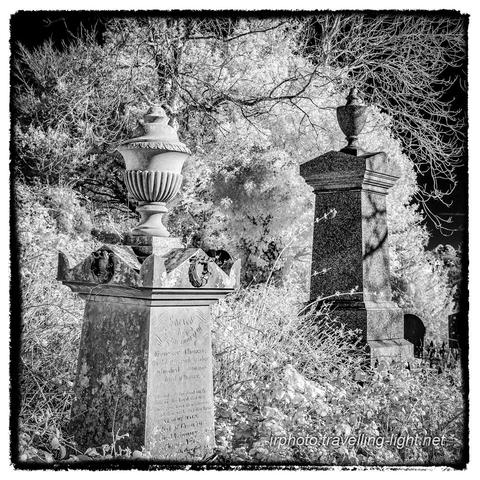 A square black and white infrared photo showing a two memorials in the form of tapered stone pillars with urns on the top, in an overgrown churchyard.