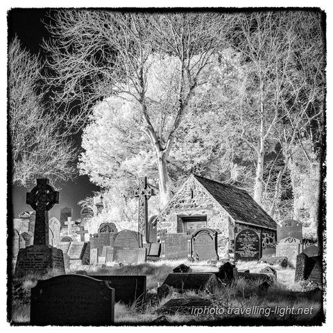 A square black and white infrared photo showing a a number of gravestones and two tall Celtic crosses in an overgrown churchyard, with a small stone building and trees behind, some with bare branches and some with leaves.