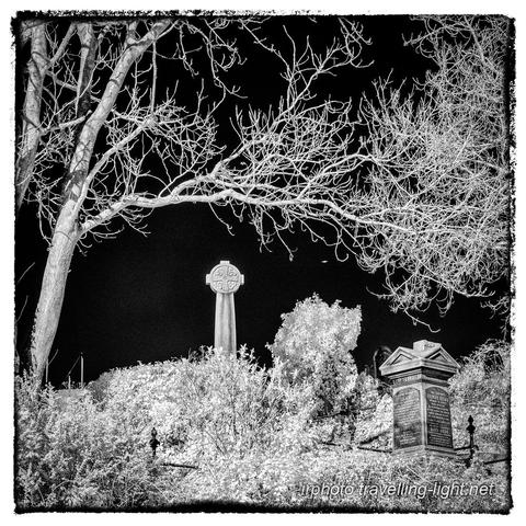 A square black and white infrared photo showing an overgrown churchyard with a memorial in the form of a square pillar and a tall Celtic cross, framed by a tree with bare branches.