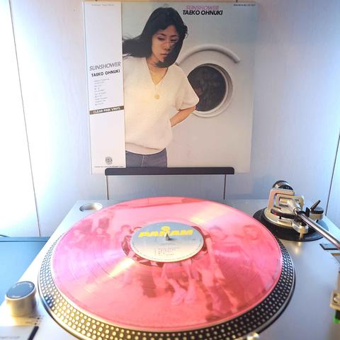 A transparent pink vinyl record sits on a turntable. Behind the turntable, a vinyl album outer sleeve is displayed. The front cover shows Taeko Ohnuki standing in front of a wall with a small circular window that has a plant inside.