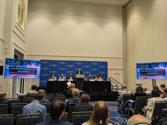 Five astronomers and an emcee on a riser in front of a blue AAS-branded background.