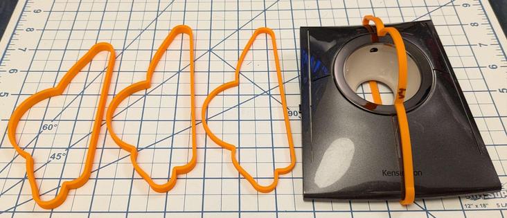 A series of orange 3d printed rings that approximate the y/z plane of the pictured trackball mouse (kensington slimblade pro) that is pictured without its red ball. The rings are on a background of a blue lines on white background cutting mat with a grid of inch markings and some diagonal lines to denote angles. The objects are laid on a plane that  tilts away from the camera and recedes slightly towards the top of the image. From left to right, the four irregularly shaped orange rings more closely approximate the shape of the center (sagittal) plane of the mouse. The fourth ring is vertical to demonstrate that it fits the roughly rectangular (tapered towards the back edge) grey shiny mouse body reasonably well.