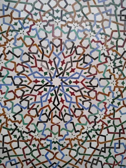 A photo of a tiling that is mainly white with colourful interlaced lines. The centre of the pattern is a ten-pointed star. The colours of the lines are light blue, black, light and dark green, orange and red. In some spaces between the lines are ornaments that look like stylised flowers or leaves.