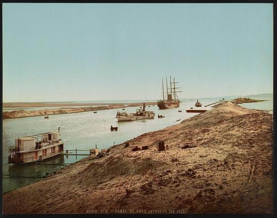The image depicts a panoramic view of the Suez Canal with several vessels navigating its waters. The foreground shows a dry, barren embankment leading up to the canal's entrance, where a ship is moored close to shore. In contrast, multiple boats and ships are scattered across various positions within the waterway, some near docks or causeways that extend into the sea.

In the background, calm seas meet distant landforms under a clear sky. A prominent feature in this landscape is an old sailing vessel with tall masts docked at one of the canal's terminals, suggesting historical significance to maritime activities around this region. The overall composition evokes a sense of early 20th-century commerce and travel within this iconic waterway leading from the Red Sea into Lake Suez (now known as Lake Timsah) in Egypt.

Notably, there is text at the bottom right corner that reads "Canaal de Suez, entrée au Lac Salé," which translates to "Suez Canal, entrance to Lake Salt" and indicates the canal's geographical significance. The image appears aged with a sepia-toned hue, likely due to its historical origin as an early 20th-century photochrome print from Photoglob Company in Zürich.

This photograph captures not only the physical aspects of the Suez Canal but also hints at the bustling economic life that once thrived along this crucial waterway.