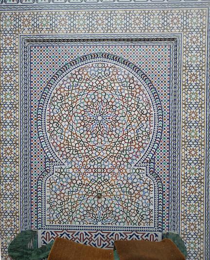 A photo of a Moroccan fountain. The tiling on the wall is mainly white with colourful interlaced lines and other patterns. The centre of the pattern is a circular disc with a rectangular portion below. Tis is surrounded by a rectangle and borders with different patterns. The colours used are light blue, black, light and dark green, orange and red.
