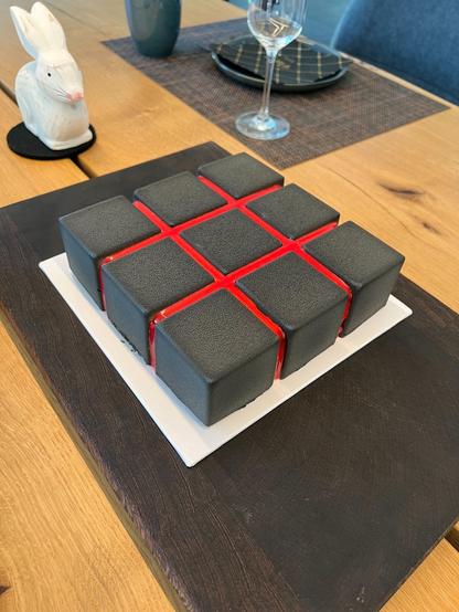Square cake (comprised of 9 equally sized cubes, like the upper row of a Rubik's Cube) with black velvet coating and red mirror glaze streaks, on a white cake board, on a dark wooden cutting board