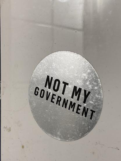 Ein silberfarbener, runder Sticker mit schwarzer Schrift. Er klebt auf weißen Fliesen. Auf dem Sticker steht in Großbuchstaben NOT MY GOVERNMENT.