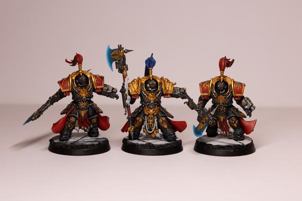 Three tall armoured models in chunky black armour with gold trim. They armour has red accents, helmet plumes and capes, with the captain in the middle having a blue helmet plume. They are all carrying blue glowing axes with a gun on the end. They are all stood on marble bases