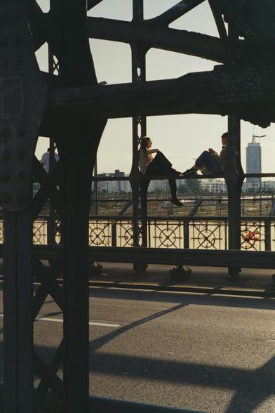 Two persons sitting on the frame/beam of a bridge talking, some high buildings on the background.
