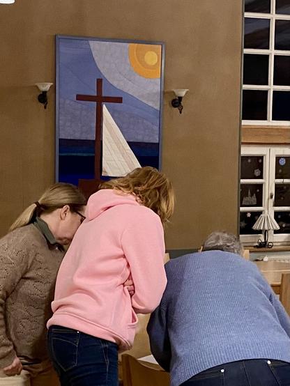 Drei Personen sind um einen Tisch versammelt und untersuchen etwas genau. Ein farbenfrohes Gemälde mit einem Segelboot, einem Kreuz und einer Sonne ist an der Wand dahinter zu sehen. Die Einstellung scheint drinnen zu sein, mit Licht kommt aus einem nahe gelegenen Fenster.