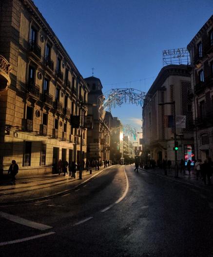Vista de la calle Recogidas desde Puerta Real. El último sol del día se refleja en los edificios y en las luces navideñas, que aún no se ha retirado. A la derecha se puede ver el edificio del antiguo cine Aliatar. Hay algunas personas caminando por la calle.
