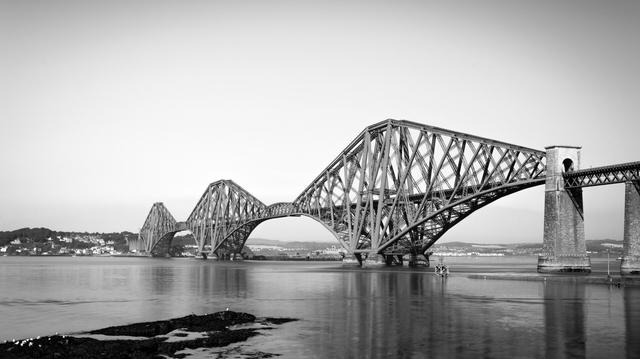 EN: A black and white view of the iconic Forth Rail Bridge in Scotland. You can see the bridge, the calm waters of the Firth of Forth and gulls on some rocks.

DE: Eine Schwarzweiß-Ansicht der berühmten Forth Rail Bridge in Schottland. Zu sehen sind die Brücke, das ruhige Wasser des Firth of Forth und einige Möwen auf Steinen im Wasser.