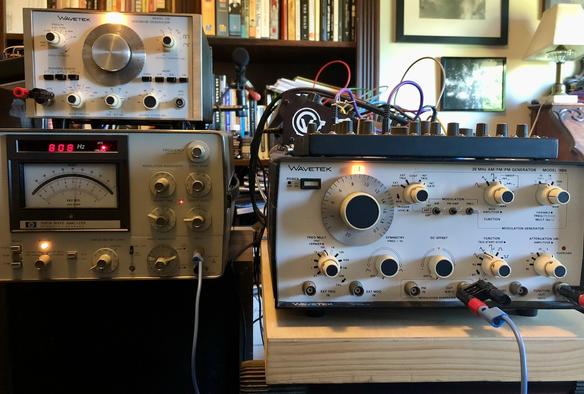 Three pieces of old school 1970s laboratory test equipment sitting in my music studio. On the left, one with a large chrome dial is sitting on top of another which has an analog meter to measure voltage or decibels. On the right and closer to you is the third piece, which has lots of dials and switches. A cable is running from it to the unit with the analog meter.