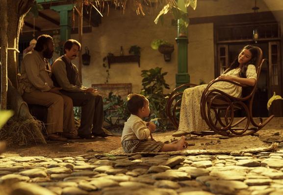 Fotograma de la serie donde se ve a un niño sentado en un suelo empedrado frente a una casa, con dos hombres sentados a su izquierda en un banco, y una mujer en una mecedora.