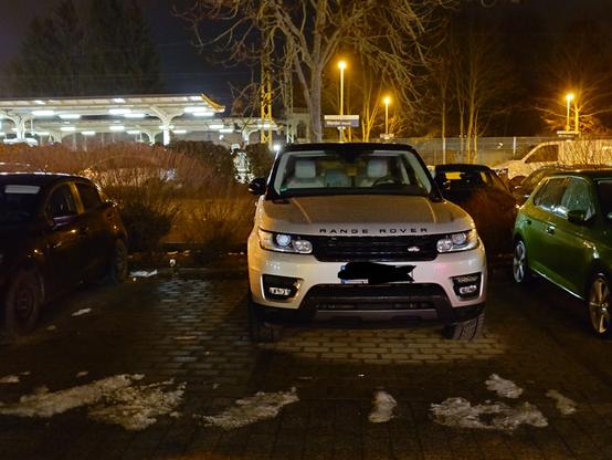 A big SUV is so broad that it takes up more than one parking space. The scene is after sunset, in the parking lot of a train station.