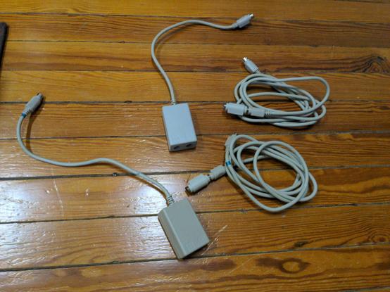 Lying on a wood floor: Two gray cables with mini-DIN connectors on them, each with a gender changer on one end. Two gray plastic boxes, each with a cord coming out of one end ending in a mini-DIN connector. One one of them you can see two mini-DIN ports opposite the cable.