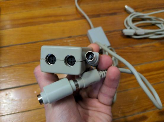 A closeup of a hand holding a LocalTalk transceiver and cable. The female ports on the transceiver are mini-DIN with three pins and a central key. One male end of the cable is visible. The other end is plugged into a gender changer.