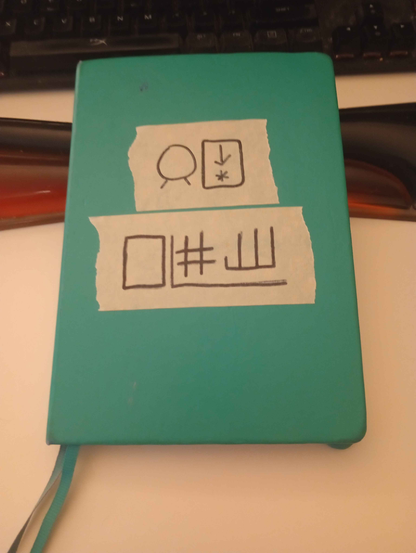 a blue-green journal with symbols written on masking tape. The symbols read, in toki pona, "jan [ni kin]. lipu pi nanpa 0111", which means "Niki, book 7".