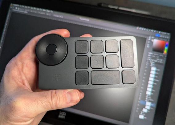 A hand holds a black XP-Pen shortcut remote with multiple buttons, in front of a graphics tablet displaying a digital editing software interface.