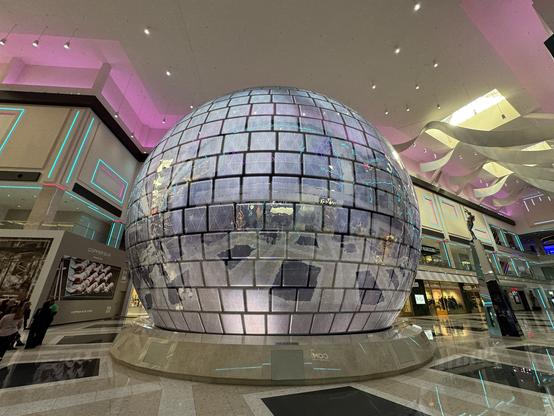A large, spherical structure covered in mirrored panels stands in a spacious shopping area, reflecting ambient light and surroundings. The interior features modern architectural elements with vibrant lighting.