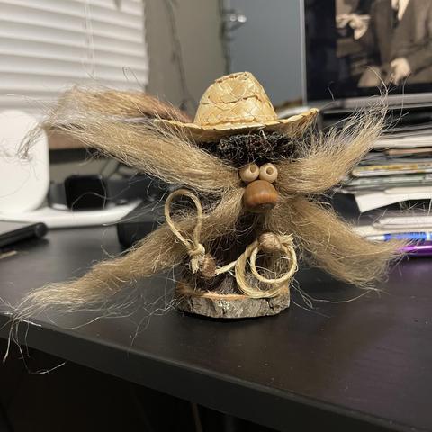 A whimsical handcrafted figure with a large mustache made of straw, wooden beads for eyes and nose, and a straw hat. It holds a small rope, sitting on a piece of wood. The background features a cluttered desk with various items.
