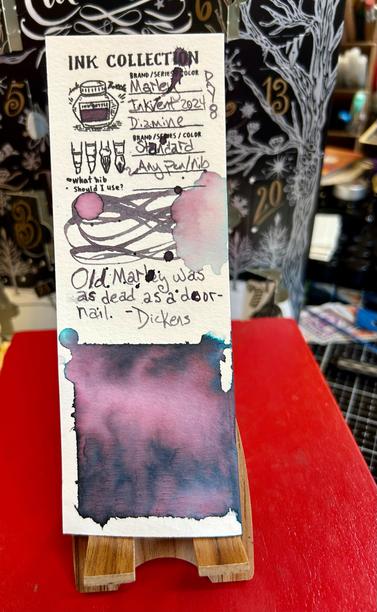 A narrow strip of watercolor paper  with an ink swatch is on a small easel that is sitting atop a red book with an advent calendar filled with ink  made by Diamine is in the background. The calendar is black with gold and silver trees and numbers with fancy lettering in white.