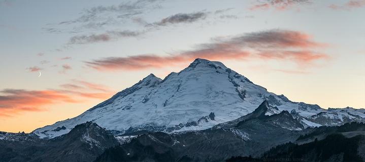 Photograph with some sunset light on clouds above Mount Baker - a volcano in Washington State. Thin slice of the moon also visible on the left side.