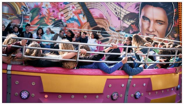 The image shows a group of people, mostly children, enjoying a ride in an amusement park. They are seated in a ride with a ring of seats that spins, a typical park attraction designed to create a thrilling experience. The people look excited and engaged, with some raising their hands in the air, a common gesture indicating thrill and enjoyment during such rides. Groups of girls interlock grips on the bars.  Boys loosely grip on, if at all.  One girl is standing and leaning towards the axis. The background features brightly colored and vibrant artistic murals, likely part of the ride’s decoration, which adds to the festive and fun atmosphere of the setting.