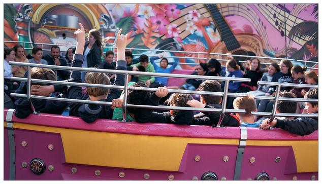 The image shows a group of people, mostly children, enjoying a ride in an amusement park. They are seated in a ride with a ring of seats that spins, a typical park attraction designed to create a thrilling experience. The people look excited and engaged, with some raising their hands in the air, a common gesture indicating thrill and enjoyment during such rides. Groups of girls interlock grips on the bars.  Boys loosely grip on, if at all.  One girl is standing and leaning towards the axis. The background features brightly colored and vibrant artistic murals, likely part of the ride’s decoration, which adds to the festive and fun atmosphere of the setting.