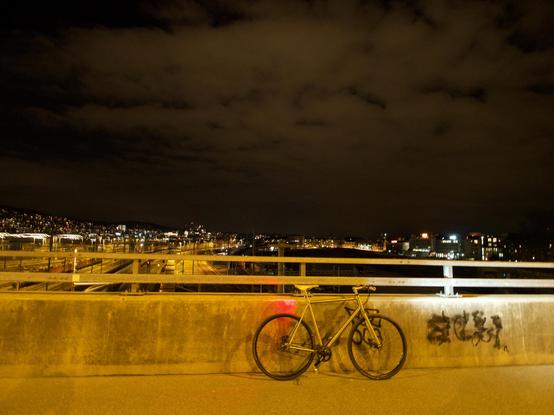 Das Bild zeigt ein Fahrrad, das nachts auf einer Brücke abgestellt wurde. Im Hintergrund sind Lichter einer Stadt sichtbar, unter einem wolkenverhangenen Himmel.