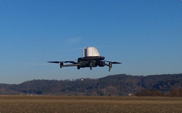 A gray dji Mavic 3E multicopter with a white 3d printed payload (PARASITE) on top, hovering over a field, a hillscape in the background, blue sky