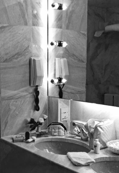 A black-and-white image of a bathroom sink area featuring marble walls, two circular sink basins, and various toiletries. There are light fixtures mounted on a mirror, a phone on the wall, a small vase with flowers, and a towel on the vanity.