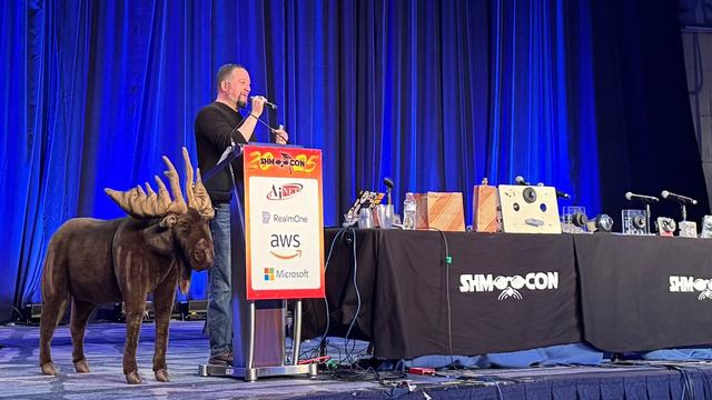 A photo of a handsome man on stage behind a podium with a moose next to him on one side and a table with locks and other items on it on the other side. He is speaking into a microphone animatedly.