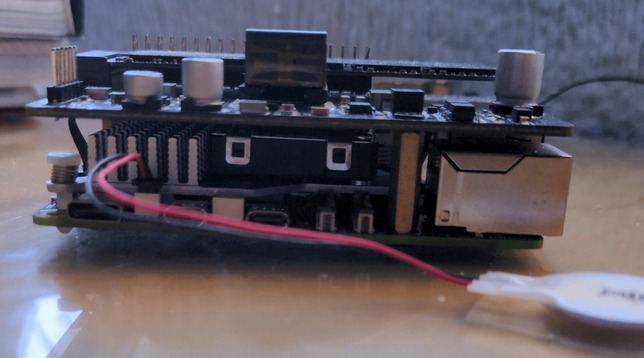 Side view of a Raspberry Pi 5 with an NVMe SSD HAT installed. The stacked design is visible, with heat sinks on the Pi for cooling, exposed connectors, and a cable leading to a small external battery connected to the RPI5, for maintaining the real-time clock. The setup remains compact and functional.