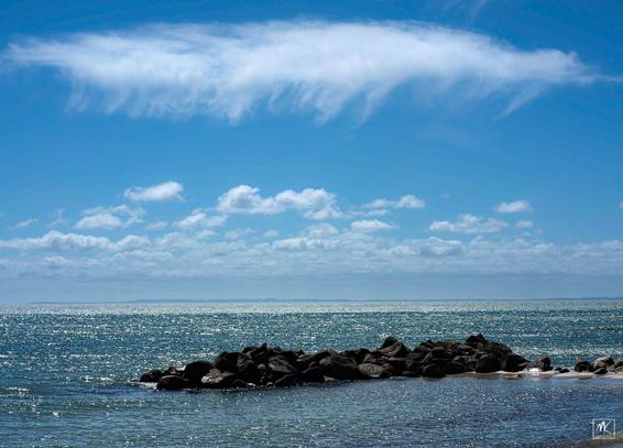 Color photo of a long pile of offshore rocks in a brightly sunlit sea with a white cloud above it and more clouds in the distance.