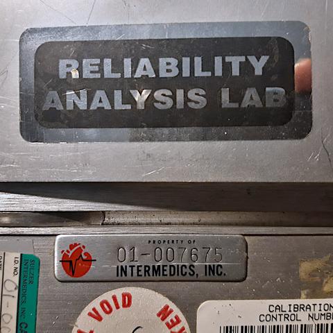 Various stickers on a piece of electronic equipment. A large sticker says 'Reliability Analysis Lab'. A smaller sticker says 'Intermedics, Inc.' and has a logo representing a heart with an electrocardiogram waveform superimposed.