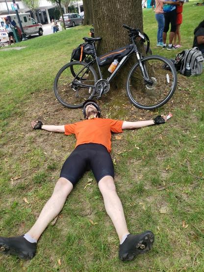 An exhausted human laying star fished on the ground. They’re wearing an orange shirt and biking gear. Their black bike with some packs is resting on a tree in the background