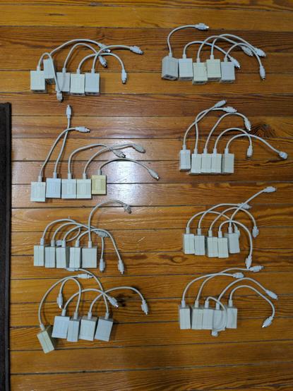 Forty PhoneNET transceivers (small gray ridged boxes with a short mini-DIN cable on one end and two phone jacks on the other) arrayed on a wood floor in groups of five. Some are faded or yellowed.