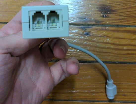 A closeup of a Caucasian hand holding a PhoneNET transceiver to present the phone jacks to the viewer. Four contacts are clearly visible in both of them and the male min-DIN plug can be seen in the background.