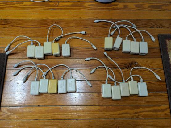 Four groups of five Farallon PhoneNET Plus transceivers (small gray boxes with rounded corners, two phone jacks on one side, and a short cable ending in a male mini-DIN connector) on a wood floor. Some are yellowed and the Farallon logotype can be made out through the glare on the shiny plastic.