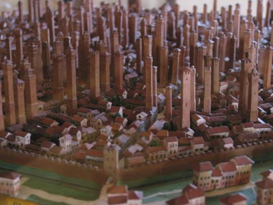 Photo of a model of Medieval Bologna, with so many earthy pink tall skinny towers sticking up from every block of the terra-cotta-roofed town that it looks like plant seeds starting to come up in spring. Around the edge you can see the city's moat and battlemented walls, looking tiny compared to the towers which rise to six or seven times the height of the three-story buildings around them.