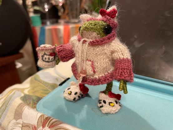 A miniature knitted green frog in a cream hoodie with dark pink trim, cat ears, and a red bow on top is holding a very small Hello Kitty mug with sculpted coffee and froth on top. She’s wearing cream slippers with cat ears, red bows, and Hello Kitty’s face embroidered on them.