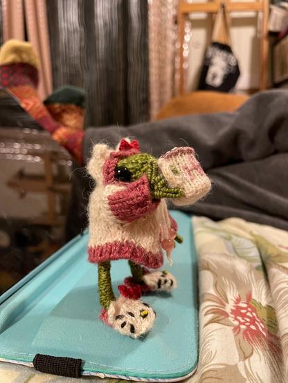 A miniature knitted green frog in a cream hoodie with dark pink trim, cat ears, and a red bow on top is sipping from a very small Hello Kitty mug with sculpted coffee and froth on top. She’s wearing cream slippers with cat ears, red bows, and Hello Kitty’s face embroidered on them.