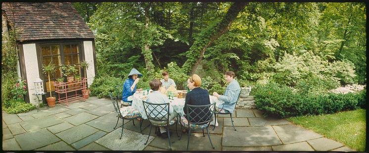 an image of a group of people eating outdoors around a table and some greenery in the background with another person standing behind them.